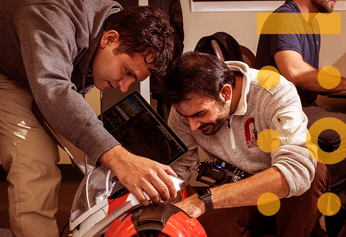 Photograph of two adults are working with their hands on an electronics project. A third student works on a computer behind them. Yellow shapes are overlaid into the photo.