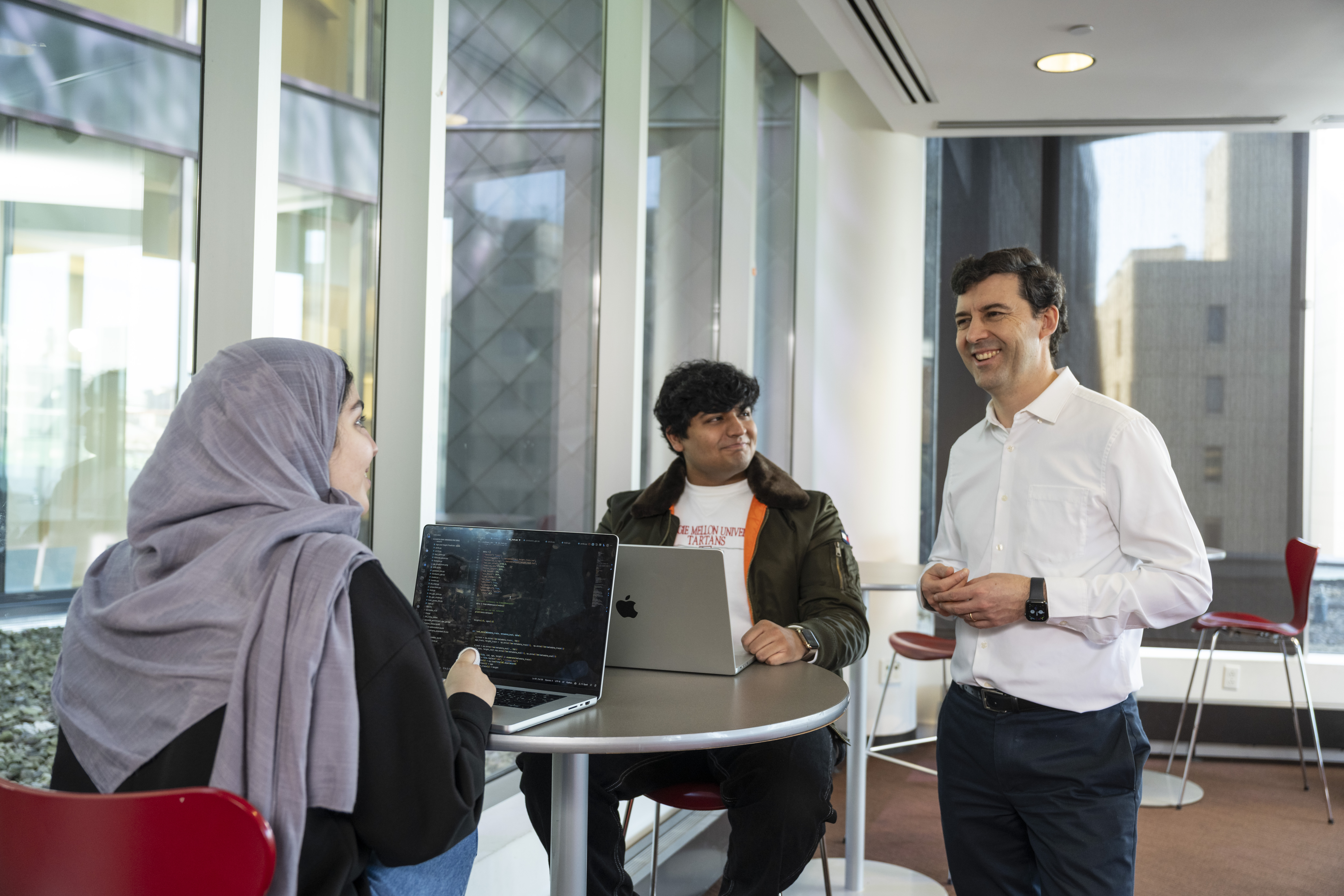 Two LTI students, a man and a woman, sit at a high-top table looking at a faculty member standing next to the table and smiling