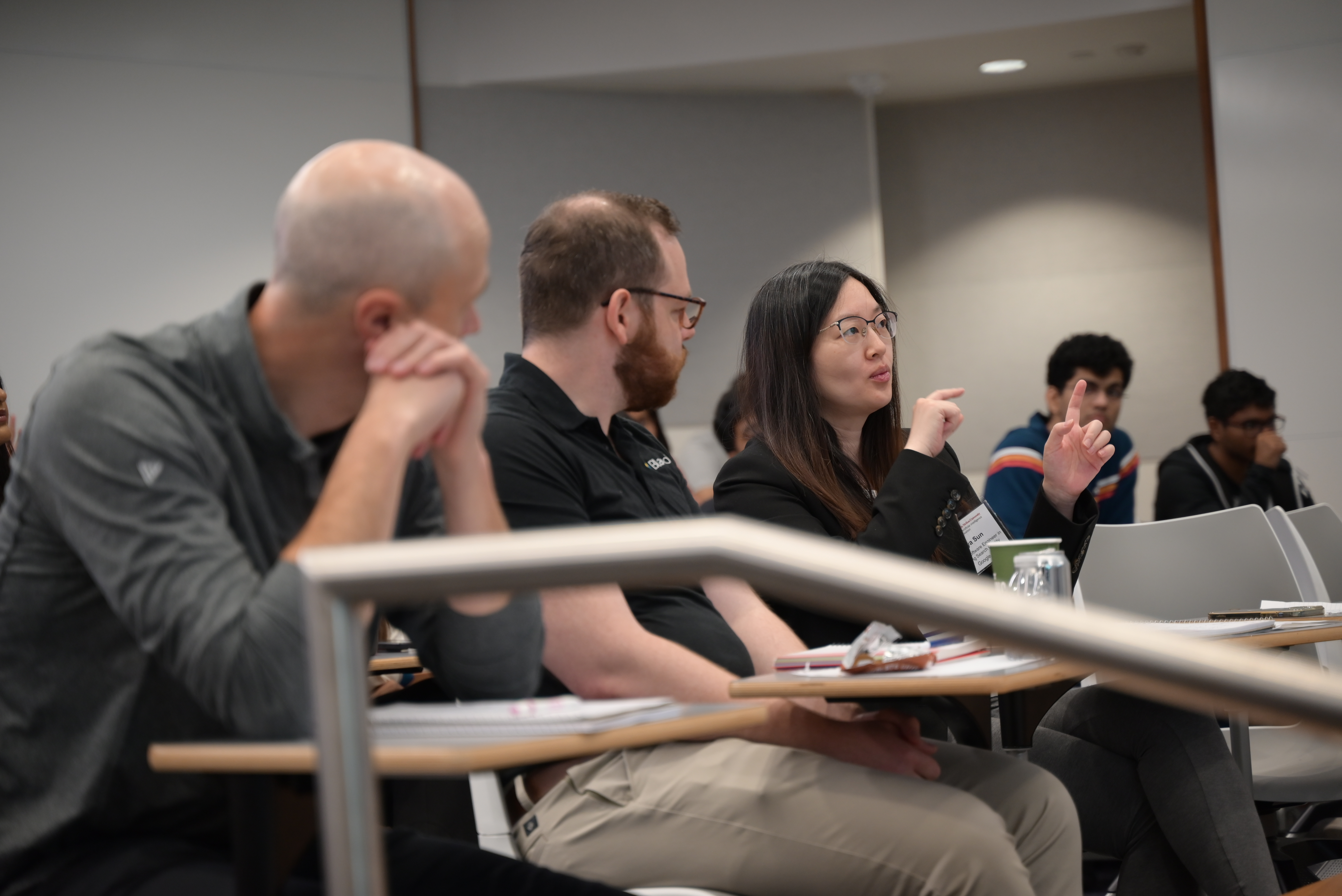 Three people seated at a table, two men and a woman, the men look on as the woman speaks